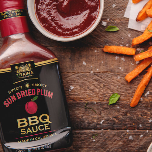 Overhead view of a textured brown wood board featuring a bottle of Sun Dried Plum BBQ Sauce, a small tub showing the sauce's thick texture, and scattered sweet potato fries, suggesting use as a delicious topping.